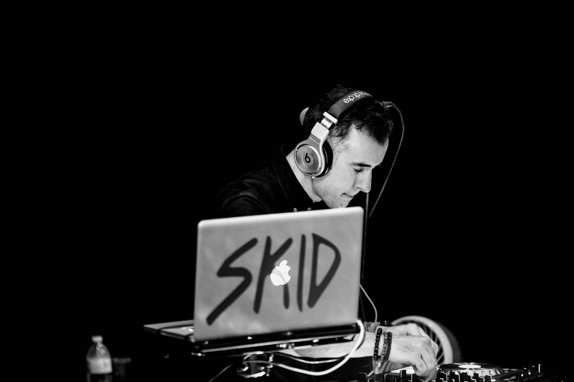 DJ wearing headphones performing with a laptop labeled SKID in a dark setting.